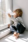 Side view of blonde little child in casual clothes playing with paper airplane while sitting barefoot on window sill on sunny day — Stock Photo