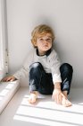 Annoyed blond little boy looking at camera with dissatisfaction while sitting on sill leaning on white wall — Stock Photo
