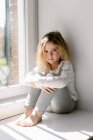 Menina irritada com cabelos encaracolados vestindo pijama cinza aconchegante olhando para a câmera com insatisfação enquanto sentado no peitoril com braços cruzados — Fotografia de Stock