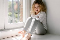 Анонімна маленька дівчинка з кучерявими волоссям у сірій затишній піжамі дивиться на камеру з незадоволенням сидячи на підвіконні з схрещеними руками — стокове фото