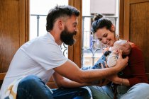 Barbuto padre e madre felice comunicare con bambino carino mentre seduto sul pavimento vicino alla finestra a casa — Foto stock