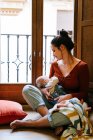 Happy mother breastfeeding baby near window — Stock Photo