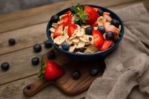 Von oben köstliche Frühstücksschüssel mit Cornflakes mit Erdbeeren und Blaubeeren auf Schneidebrett gelegt und mit Leinentuch und Beeren um Schüssel auf Holzgrund dekoriert — Stockfoto