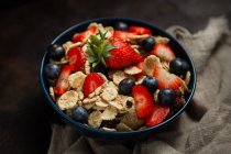 Blick von oben auf köstliche Frühstücksschüssel mit Cornflakes mit Erdbeeren und Blaubeeren auf Schneidebrett gelegt und mit Leinentuch und Beeren um Schale auf Holzgrund dekoriert — Stockfoto