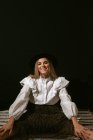 Jovem alegre mulher loira bonito em roupa elegante e chapéu sentado no chão e sorrindo para a câmera contra fundo preto — Fotografia de Stock