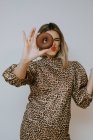 Молодая женщина в платье с леопардовым отпечатком надутых губ и глядя в камеру через сладкий шоколадный пончик на сером фоне — стоковое фото