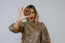 Young cute blonde female in dress with leopard print pouting lips and looking at camera through sweet chocolate doughnut against gray background — Stock Photo