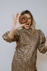 Молода жінка в одязі з леопардовим принтом, розглядаючи камеру через солодкий шоколадний пончик на сірому фоні — стокове фото