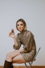 Glückliche junge blonde Frau im trendigen Kleid mit Leopardenmuster lächelt lächelnd in die Kamera und beißt leckeren Schokokrapfen, während sie auf einem Stuhl vor grauem Hintergrund sitzt — Stockfoto