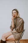 Joyeux jeune femme blonde en robe à la mode avec imprimé léopard souriant en regardant la caméra mordre beignet chocolat savoureux tout en étant assis sur une chaise sur fond de mur gris — Photo de stock