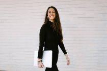 Cheerful young woman with modern laptop smiling and looking away while walking against white wall on city street — Stock Photo