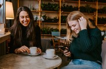 Young women laughing at joke while drinking coffee and using smartphone during meeting in cozy restaurant — Stock Photo