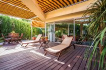 Salones de madera y mesa con sillas colocadas en la amplia terraza de la casa de campo moderna rodeada de plantas verdes - foto de stock