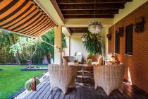 Table en osier avec chaises placées sur la terrasse de jardin spacieuse de la maison de campagne moderne entourée de plantes vertes — Photo de stock