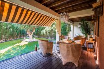 Table en bois avec chaises placées sur la grande terrasse de jardin de la maison de campagne moderne entourée de plantes vertes — Photo de stock