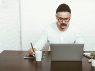 Maschio barbuto concentrato in occhiali e copricapo con netbook per lavorare a casa in quarantena — Foto stock