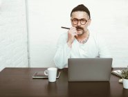 Varón barbudo concentrado en gafas y sombreros usando netbook para trabajar en casa en tiempo de cuarentena - foto de stock