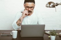 Masculin barbu concentré dans des lunettes et des coiffures à l'aide d'un netbook pour travailler à la maison pendant la quarantaine — Photo de stock