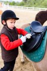 Vue latérale du petit jockey en casque de protection ajustant l'étrier sur la selle avant de monter poney asymétrique à l'école équestre — Photo de stock