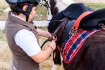 Side view of little jockey in protective helmet adjusting stirrup on saddle before riding skewbald pony in equestrian school — Stock Photo
