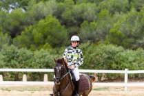 Jockey adolescente en casco mirando a la cámara mientras monta caballo marrón en la arena de doma durante el entrenamiento en la escuela ecuestre - foto de stock