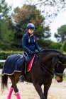 Девушка-жокей в шлеме катается на коричневой лошади под ветвями деревьев на выездной арене во время тренировки в конной школе — стоковое фото