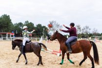 Adolescentes em capacetes jogando bola uns para os outros enquanto montam cavalos na arena de adestramento durante a aula na escola equestre — Fotografia de Stock