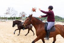 Teenager in Helmen werfen sich beim Reiten auf Pferden im Dressurviereck während des Unterrichts in der Reitschule Ball zu — Stockfoto