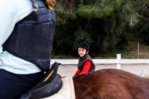 Niño en pony romano y chica adolescente anónima en caballo marrón montar a caballo en la arena doma durante la lección en la escuela ecuestre - foto de stock