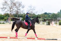 Adolescente jóquei no capacete montando cavalo marrom sob galhos de árvore na arena de curativo durante o treinamento na escola equestre — Fotografia de Stock
