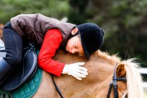 Side view of child with closed eyes in jockey costume sitting in saddle and hugging roan pony during lesson in horseback riding school — Stock Photo