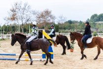 Teen Jockeys in Helmen kommunizieren miteinander, während sie gehorsame Pferde auf sandigem Dressurviereck während des Unterrichts in der Reitschule reiten — Stockfoto