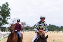 Happy teen jockeys in helmets communicating with each other while riding obedient horses on sandy dressage arena during lesson in equestrian school — Stock Photo