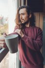 Дорослий бородатий чоловік з довгим волоссям насолоджується гарячим напоєм і дивиться у вікно, проводячи час вдома — стокове фото