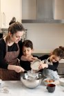 Братья и сёстры с миской муки улыбаются, помогая матери в фартуке готовить тесто на уютной кухне дома — стоковое фото