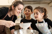 Irmãos com tigela de farinha sorrindo enquanto ajudam a mãe no avental para preparar pastelaria na cozinha acolhedora em casa — Fotografia de Stock