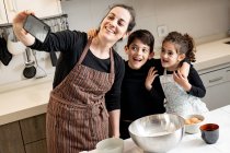 Femme heureuse dans tablier souriant et prenant selfie avec téléphone portable avec des enfants heureux tout en cuisinant la pâtisserie ensemble dans une cuisine confortable à la maison — Photo de stock