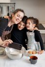 Счастливая женщина в фартуке улыбается и делает селфи с мобильным телефоном со счастливыми детьми, готовя вместе кондитерские изделия на уютной кухне дома — стоковое фото