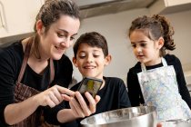 Happy woman in apron smiling and taking selfie with mobile phone with happy children while cooking pastry together in cozy kitchen at home — Stock Photo