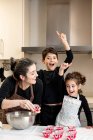 Mulher adulta feliz sorrindo e olhando para crianças animadas apontando para cima enquanto cozinham cupcakes em casa juntos — Fotografia de Stock