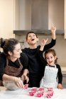 Feliz mujer adulta sonriendo y mirando a los niños emocionados apuntando hacia arriba mientras cocinan cupcakes en casa juntos - foto de stock