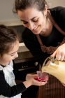 Happy woman smiling and pouring batter into paper cup while preparing cupcakes with little daughter on weekend day at home — Stock Photo