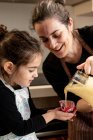 Femme heureuse souriant et verser de la pâte dans une tasse en papier tout en préparant des cupcakes avec petite fille le week-end à la maison — Photo de stock