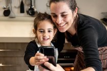 Mujer feliz en delantal sonriendo y tomando selfie con teléfono móvil con niña feliz mientras cocinan pastelería juntos en la acogedora cocina en casa - foto de stock