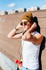 Giovane bel maschio hipster con le cuffie appoggiate al muro vicino allo skateboard prima di ascoltare musica nella giornata di sole in città — Foto stock