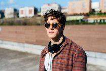 Красивый молодой человек в клетчатой рубашке и солнечных очках, смотрящий в камеру, стоя на размытом фоне городской улицы — стоковое фото