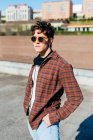 Beau jeune homme en chemise à carreaux et lunettes de soleil regardant la caméra tout en se tenant debout sur fond flou de la rue de la ville — Photo de stock
