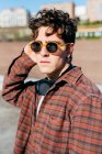 Handsome young man in checkered shirt and sunglasses looking at camera while standing on blurred background of city street — Stock Photo