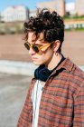 Schöner junger Mann in kariertem Hemd und Sonnenbrille, der auf verschwommenem Hintergrund der Stadtstraße in die Kamera blickt — Stockfoto
