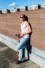 Young handsome male hipster with headphones leaning on wall near skateboard before listening to music on sunny day in city — Stock Photo